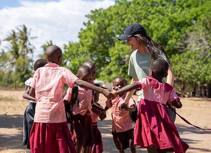 A doTERRA Healing Hands volunteer in a black cap holds hands with school-aged children in uniform, dancing in a circle.