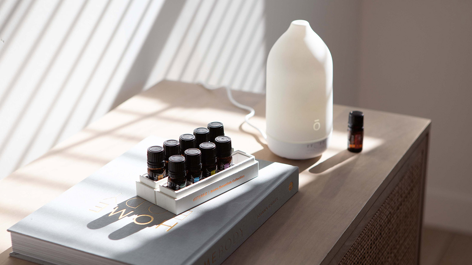 A doTERRA Laluz diffuser sits on a wooden cabinet beside a small bottle of essential oil and a tray of doTERRA oils for a homemade diffuser blend.