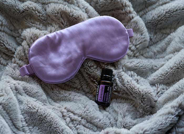 A bottle of doTERRA Lavender essential oil rests on a soft, cozy blanket beside a plush sleep mask, evoking a calm and restful bedtime atmosphere.