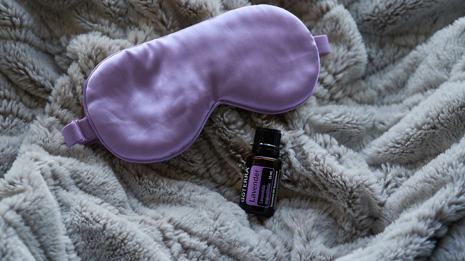 A bottle of doTERRA Lavender essential oil rests on a soft, cozy blanket beside a plush sleep mask, evoking a calm and restful bedtime atmosphere.